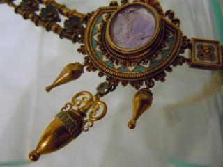 Lavendar medalion within multiple layers of gold ornament and blue and white collar, above urn pendants